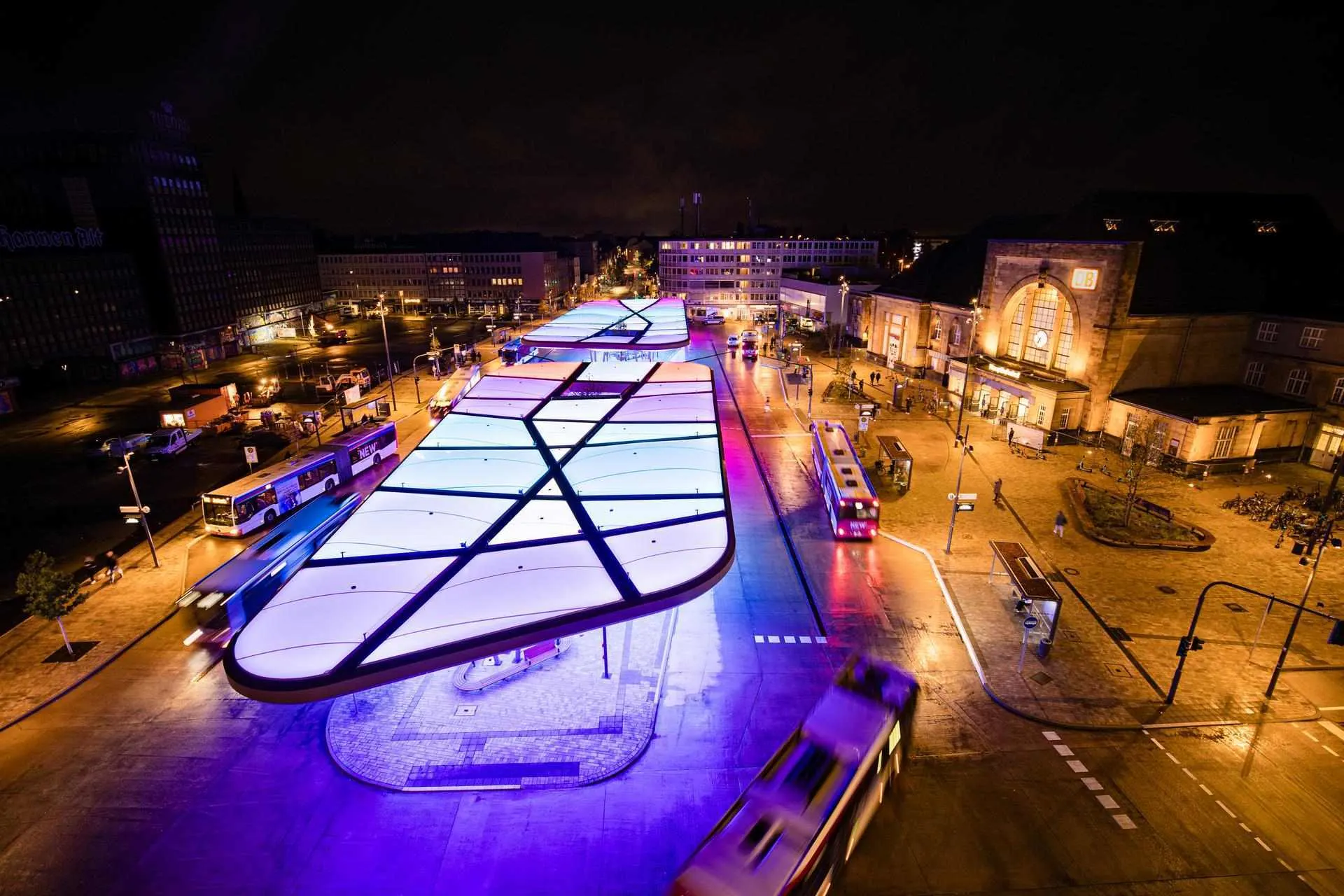 Das Bild zeigt den Zentralen Omnibusbahnhof (ZOB) in Mönchengladbach bei Nacht. Die moderne Dachkonstruktion ist farbig beleuchtet und prägt das Stadtbild. Rechts im Bild befindet sich der Hauptbahnhof, links das markante Gebäude „Haus Westland“. Die Szene wirkt dynamisch durch Lichtspuren vorbeifahrender Fahrzeuge.
