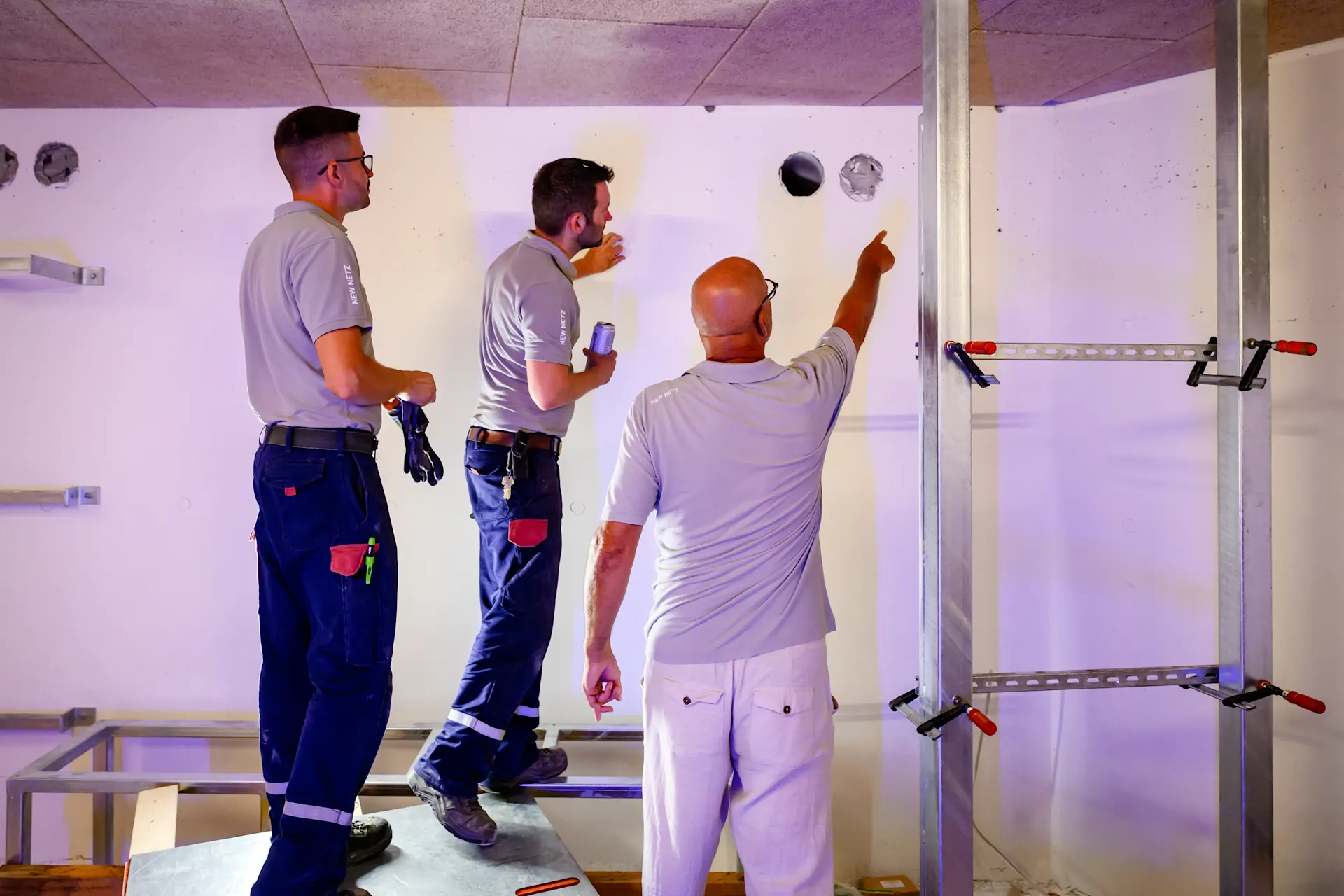 Drei Handwerker arbeiten gemeinsam an der Montage einer Trennwand in einem Innenraum. Zwei von ihnen stehen auf einem Podest und bearbeiten die Wand, während der dritte eine Metallkonstruktion mit Schraubzwingen fixiert. 