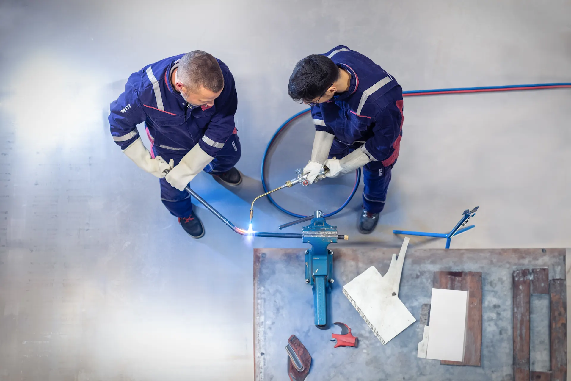 Zwei Arbeiter in blauen Schutzanzügen und Handschuhen arbeiten von oben betrachtet an einem Metallwerkstück auf dem Boden. Einer hält einen Schneidbrenner, während daneben blaue Schläuche und Werkzeuge liegen.