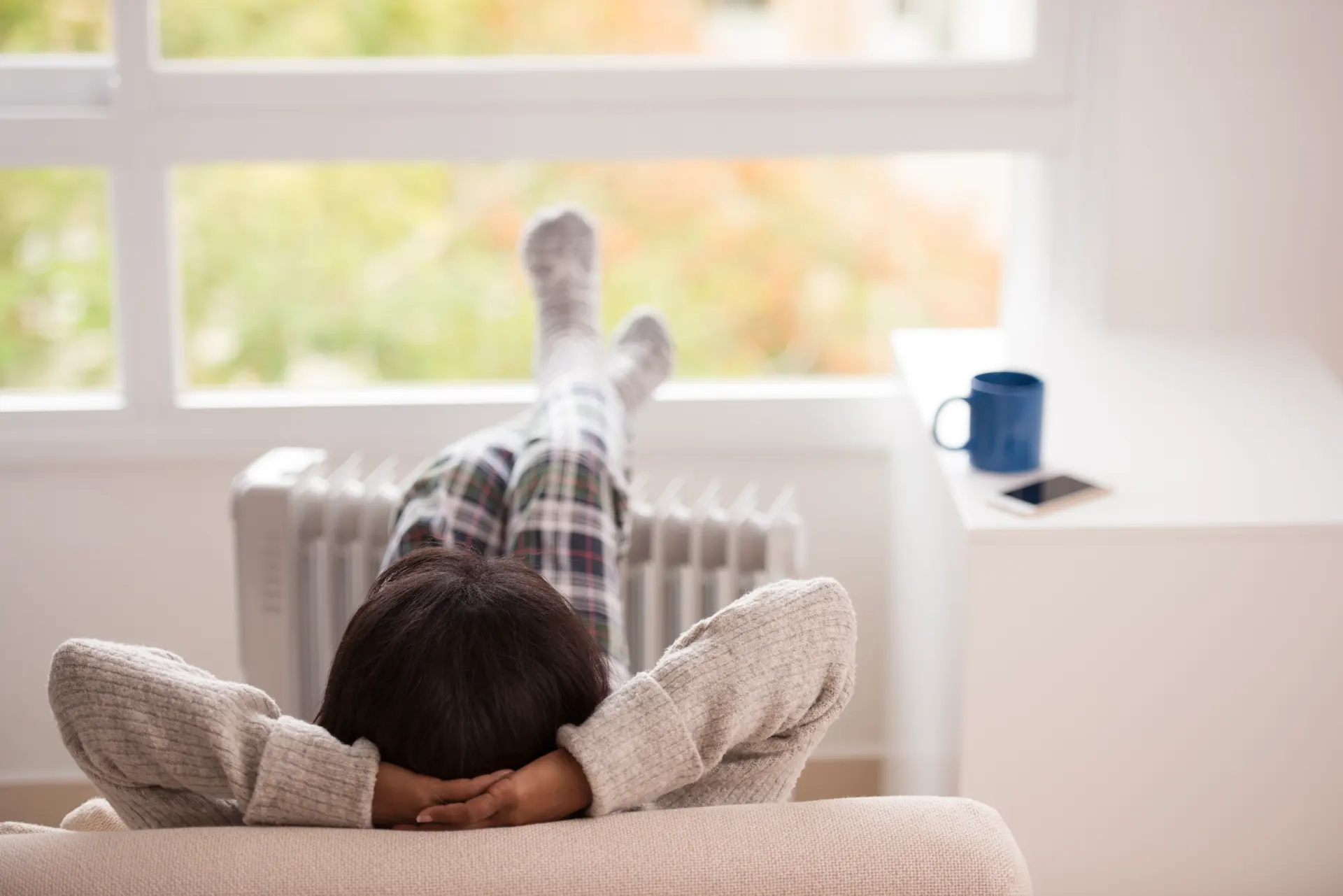Eine Person in karierter Pyjamahose und Wollsocken liegt entspannt auf einem beigen Sofa mit den Beinen nach oben an einer Heizung gelehnt. Im Hintergrund steht eine blaue Tasse auf einer Fensterbank vor einem großen Fenster. 