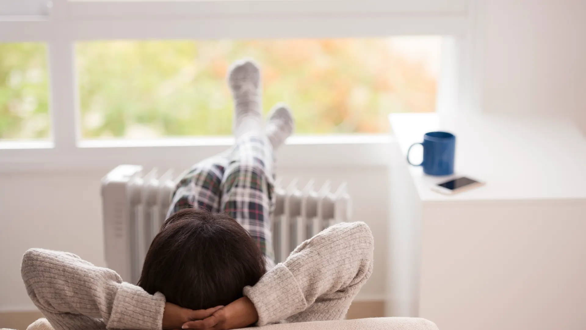 iStock-658574596 RuslanDashinsky Eine Person in karierter Pyjamahose und Wollsocken liegt entspannt auf einem beigen Sofa mit den Beinen nach oben an einer Heizung gelehnt. Im Hintergrund steht eine blaue Tasse auf einer Fensterbank vor einem großen Fenster.