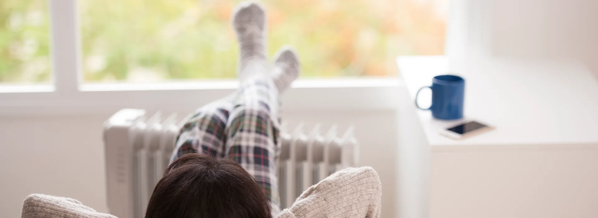 iStock-658574596 RuslanDashinsky Eine Person in karierter Pyjamahose und Wollsocken liegt entspannt auf einem beigen Sofa mit den Beinen nach oben an einer Heizung gelehnt. Im Hintergrund steht eine blaue Tasse auf einer Fensterbank vor einem großen Fenster.