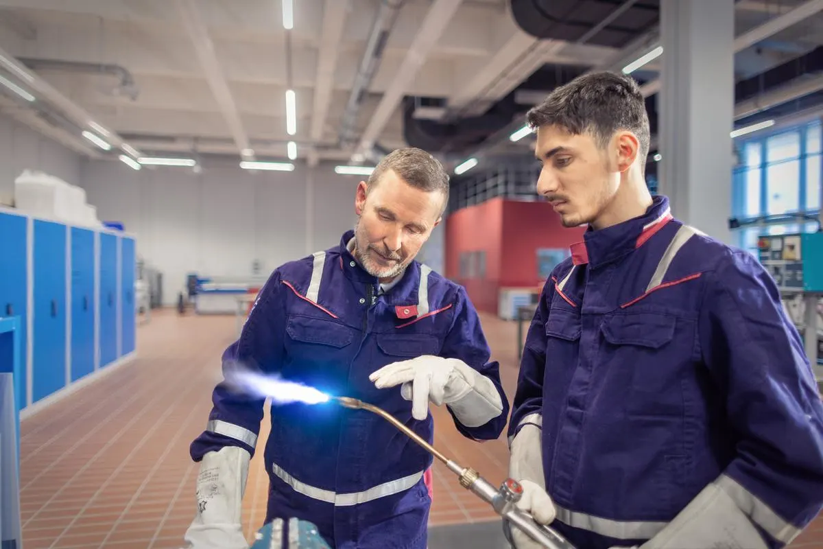 Ein Ausbilder und ein Auszubildender arbeiten gemeinsam in einer Werkhalle mit einem Schweißbrenner. Beide tragen Schutzkleidung und konzentrieren sich auf die bläuliche Flamme.