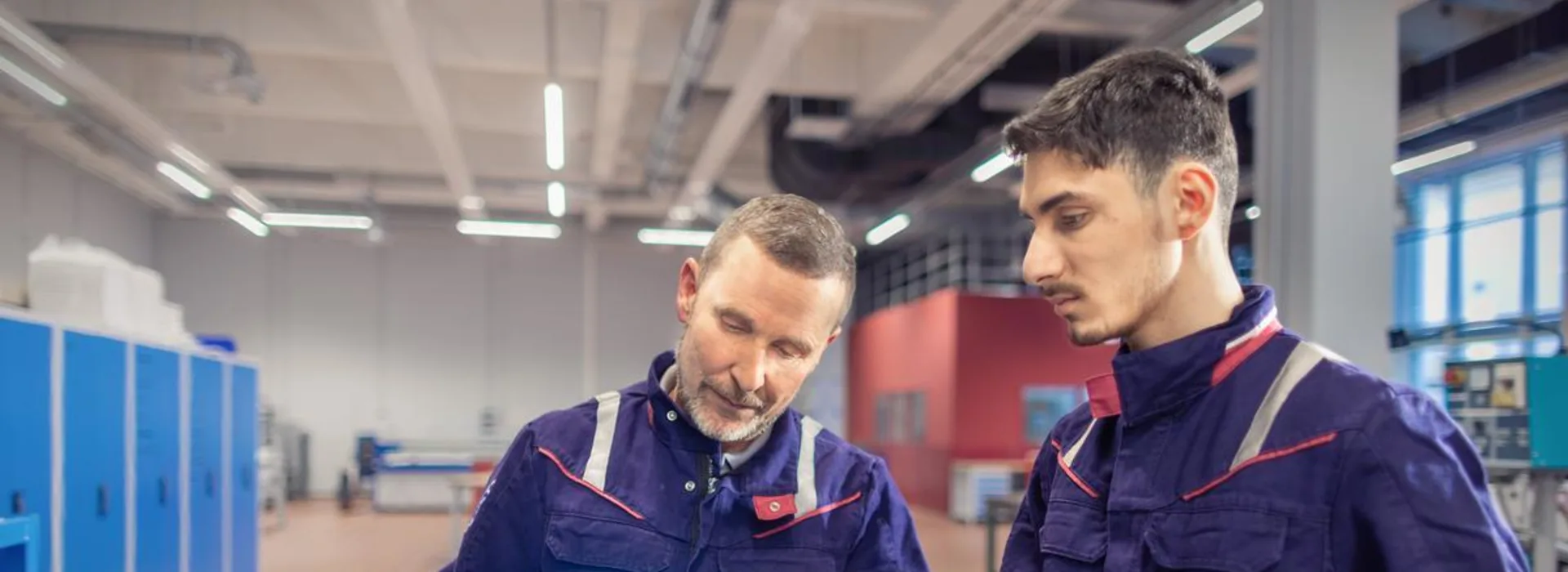 Technische Ausbildungssituation Ein Ausbilder und ein Auszubildender arbeiten gemeinsam in einer Werkhalle mit einem Schweißbrenner. Beide tragen Schutzkleidung und konzentrieren sich auf die bläuliche Flamme.