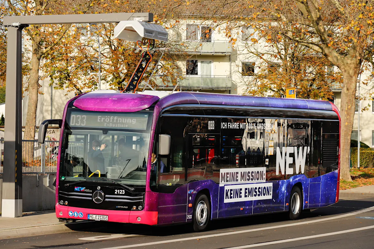 Ein moderner Elektrobus mit violett-blauer Lackierung steht an einer Haltestelle, auf der Seite ist die Werbung „MEINE MISSION: KEINE EMISSION!“ zu lesen. An der Ladestation mit Oberleitung ist der Bus zum Aufladen angeschlossen. 