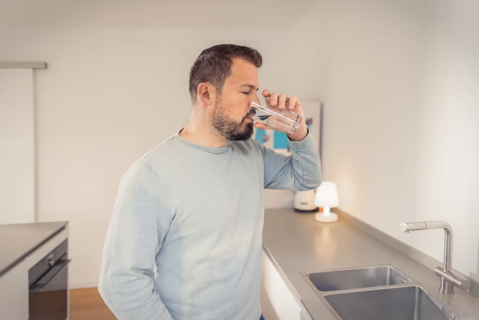 Ein Mann steht in der Küche und trinkt Leitungswasser aus einem Glas 