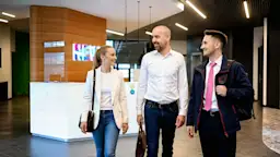 Drei Kolleg.innen am Empfang der NEW Verwaltung Drei Berufstätige in Business-Kleidung gehen lächelnd durch eine moderne Bürolobby mit hellem Holzdesign und dem bunten NEW-Logo im Hintergrund. Die Frau trägt einen beigen Blazer und Jeans, der Mann in der Mitte ein weißes Hemd, und der rechte Mann eine dunkelblaue Arbeitsjacke mit pinken Akzenten. Die Lobby verfügt über einen weißen Empfangstresen, grüne Wandelemente und moderne Deckenbeleuchtung.