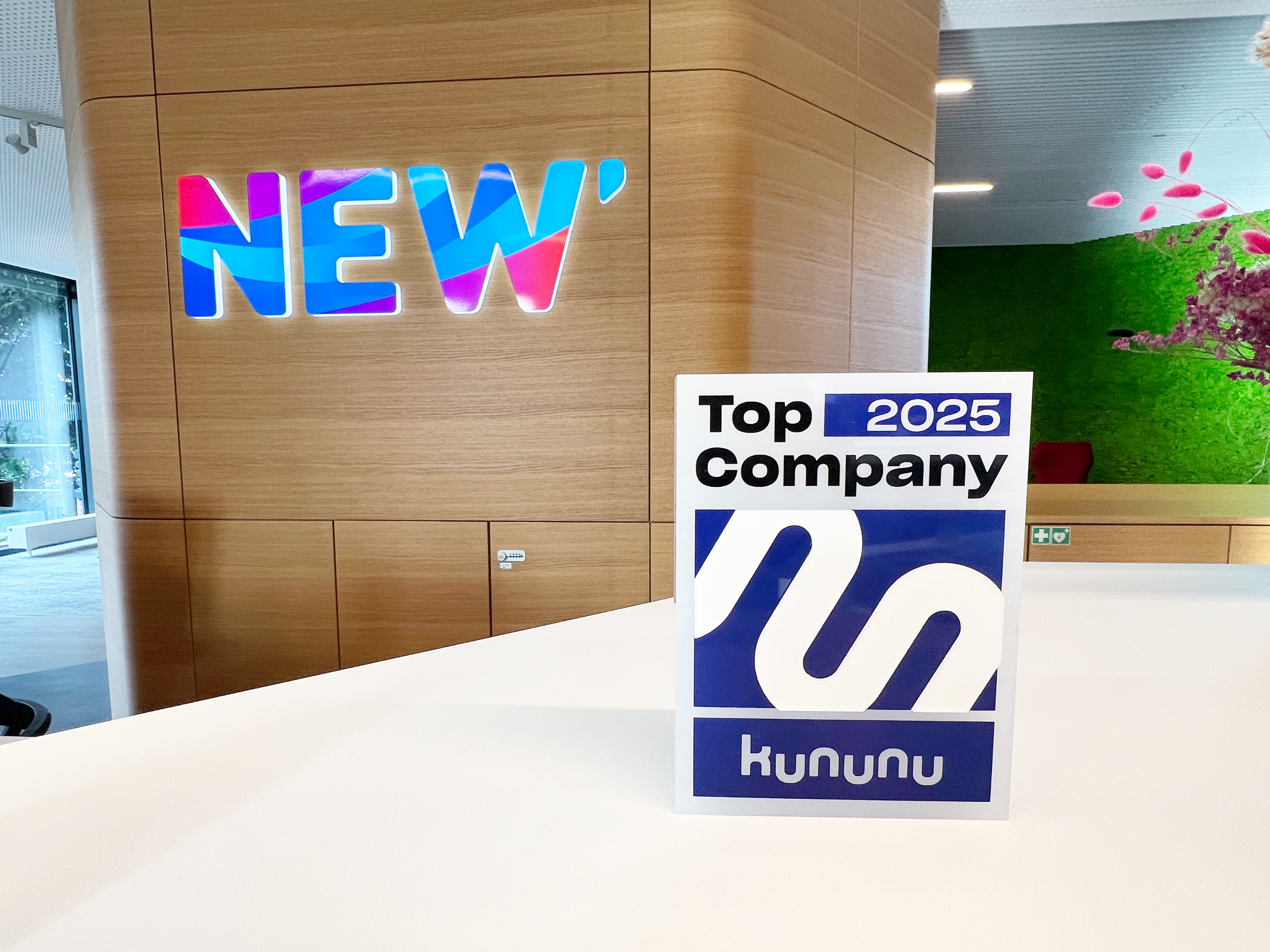 Ein moderner Empfangsbereich mit weißem Tresen, an dem eine Auszeichnung „Top Company 2025“ von kununu steht. An der Holzwand dahinter ist ein buntes, farbverlaufendes Logo mit dem Schriftzug „NEW“ angebracht.
