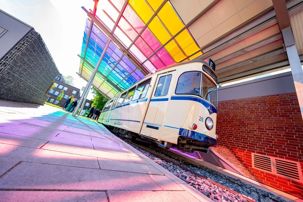 Ein restaurierter historischer Straßenbahn-Triebwagen steht unter einem modernen, farbig verglasten Dach am Gleis. Die Szene ist bei Tageslicht aufgenommen und zeigt die Bahn als Veranstaltungsort auf dem Gelände der NEW-Hauptverwaltung. 