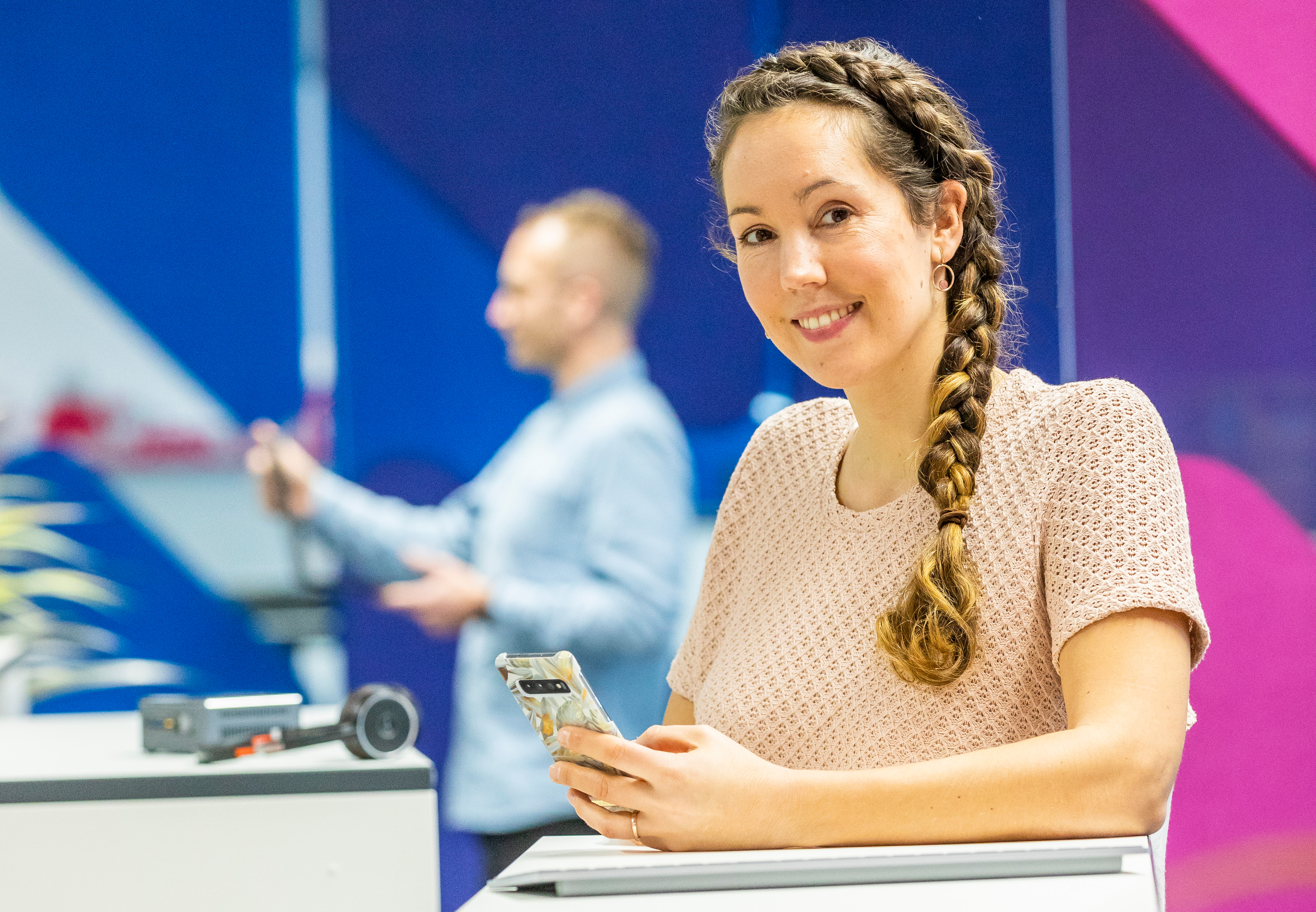 Eine junge Frau mit geflochtenem Zopf lächelt in die Kamera und hält ein Smartphone in der Hand. In der Unschärfe im Hintergrund steht ein Mann in einem modernen, farbenfrohen Büro.