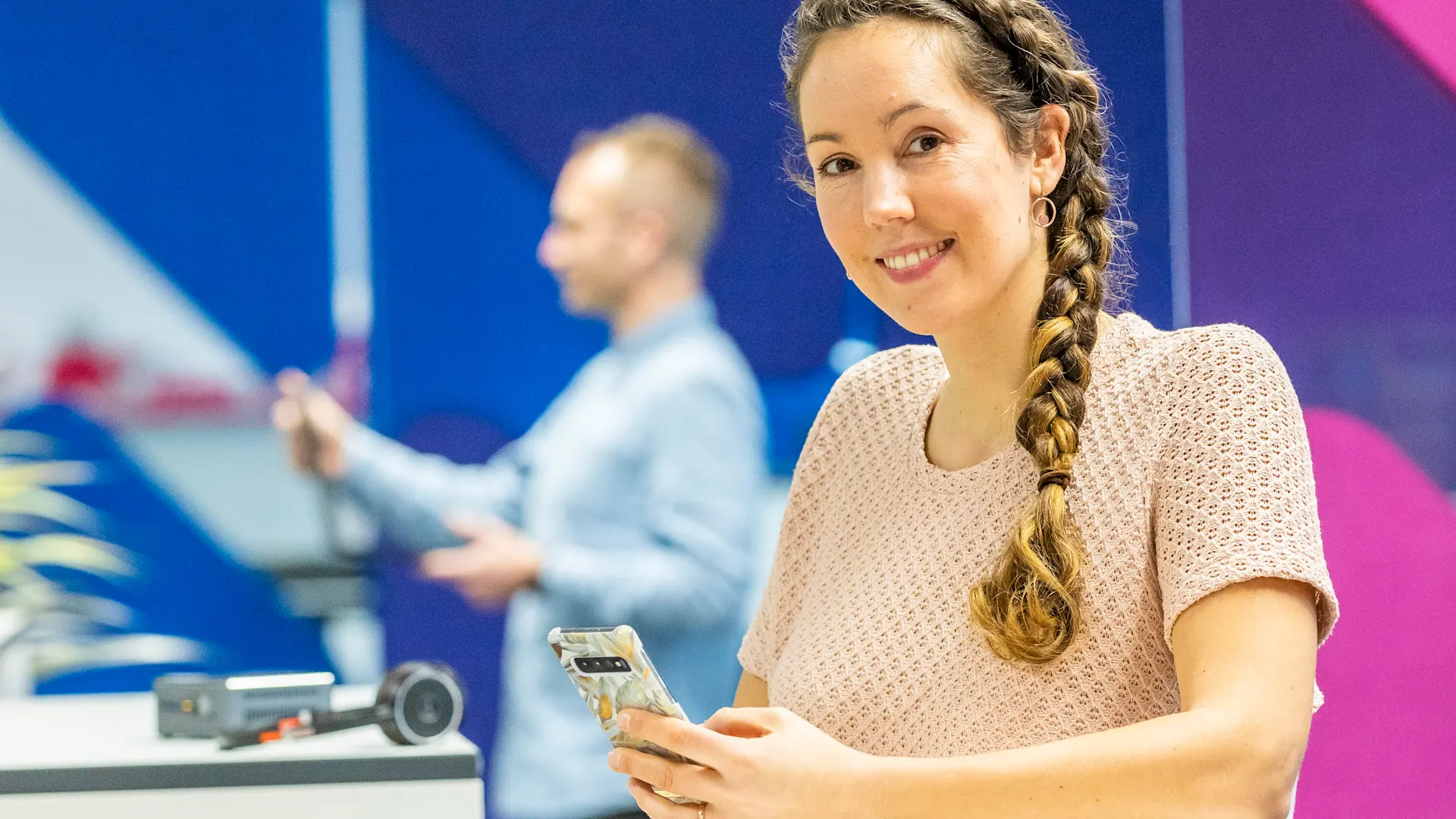 Jennifer Blasic - Digital Transfer Managerin Eine junge Frau mit geflochtenem Zopf lächelt in die Kamera und hält ein Smartphone in der Hand. In der Unschärfe im Hintergrund steht ein Mann in einem modernen, farbenfrohen Büro.