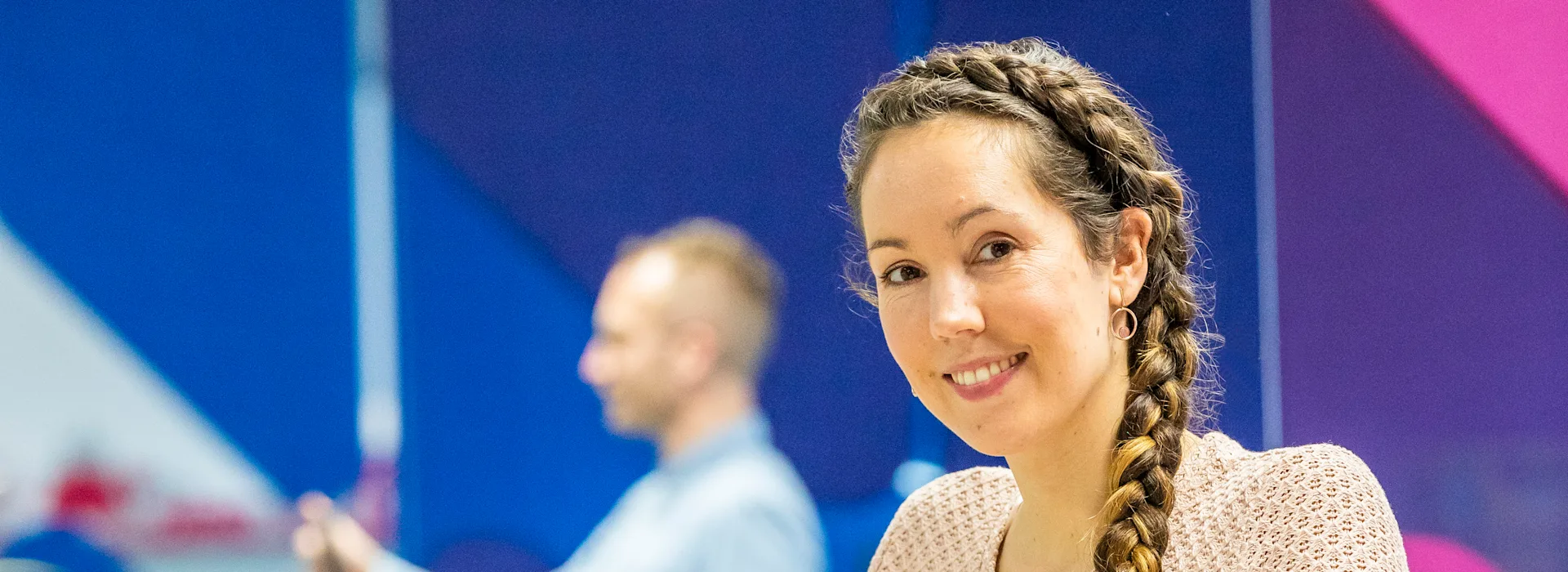 Jennifer Blasic - Digital Transfer Managerin Eine junge Frau mit geflochtenem Zopf lächelt in die Kamera und hält ein Smartphone in der Hand. In der Unschärfe im Hintergrund steht ein Mann in einem modernen, farbenfrohen Büro.