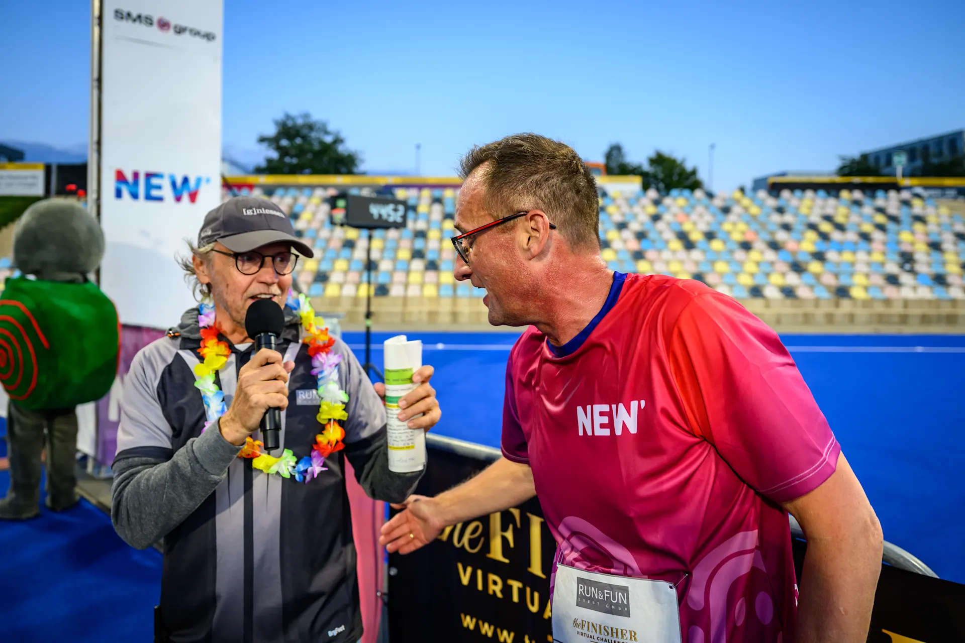 Ein Moderator mit Mikrofon und bunter Blumenkette interviewt einen Läufer in pinkem NEW-Shirt bei einer Laufveranstaltung im Stadion. Im Hintergrund sind die gelb-blauen Zuschauerränge und weitere Teilnehmer zu sehen.