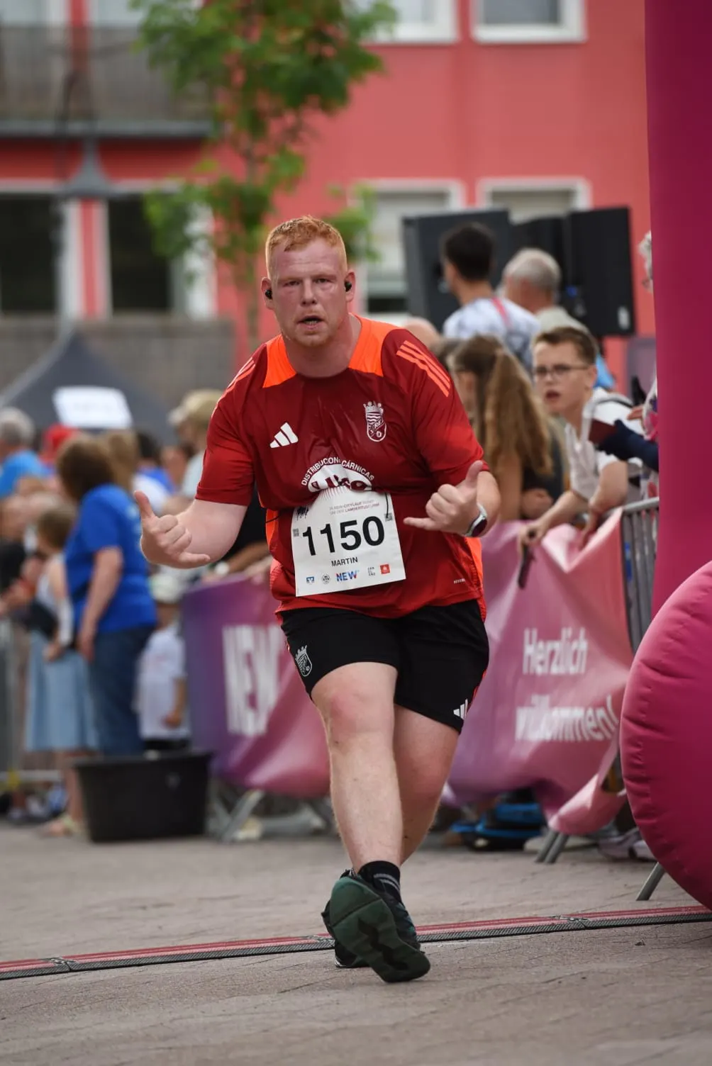 Martin Giesers-Aviles läuft mit Startnummer 1150 und entschlossenem Gesichtsausdruck in rotem Lauftrikot bei einer Laufveranstaltung, im Hintergrund sind Zuschauer:innen und ein rotes Gebäude zu sehen.