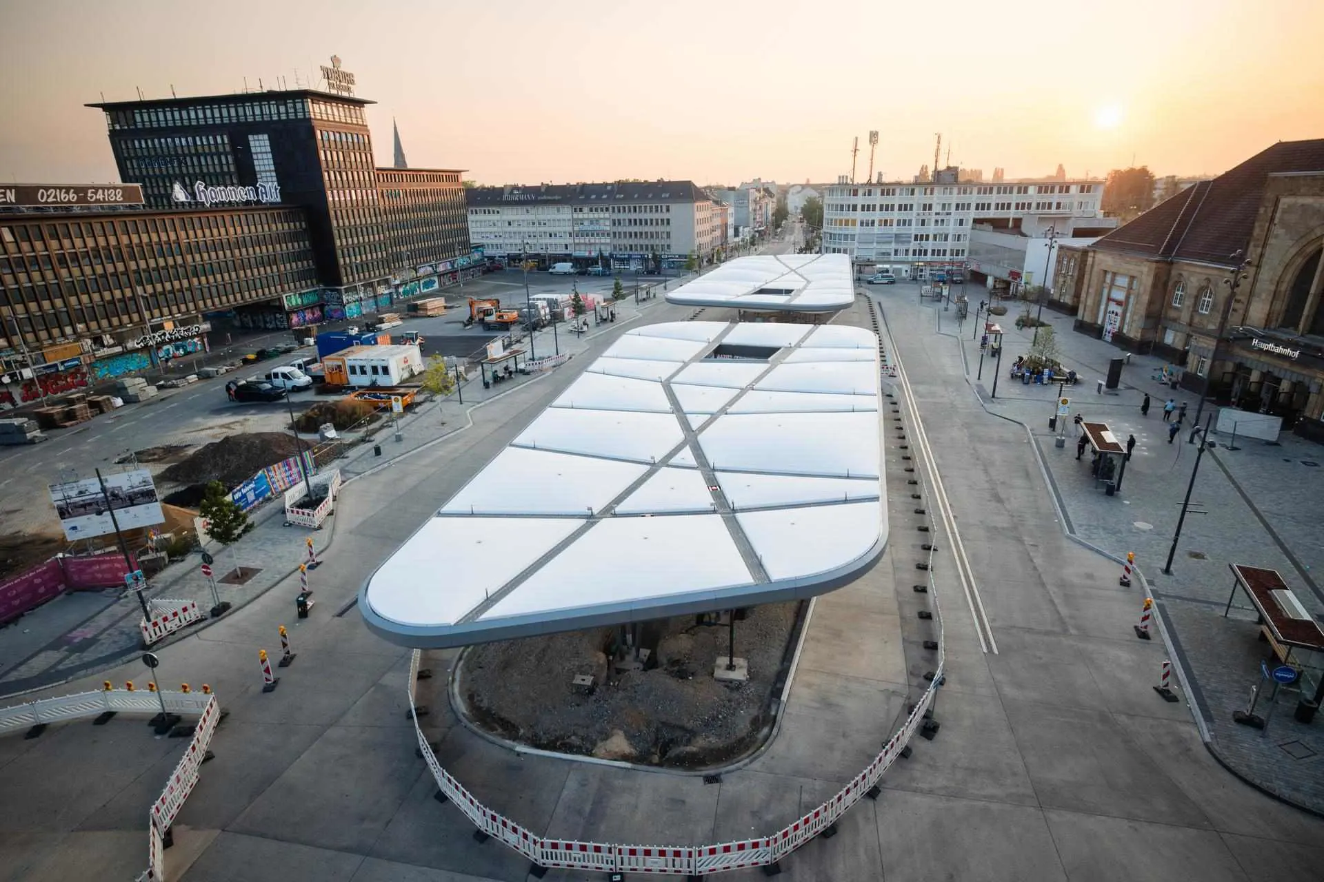 Das Bild zeigt den Zentralen Omnibusbahnhof (ZOB) in Mönchengladbach bei Sonnenaufgang. Die moderne Dachkonstruktion prägt das Stadtbild. Rechts im Bild befindet sich der Hauptbahnhof, links das markante Gebäude „Haus Westland“. 