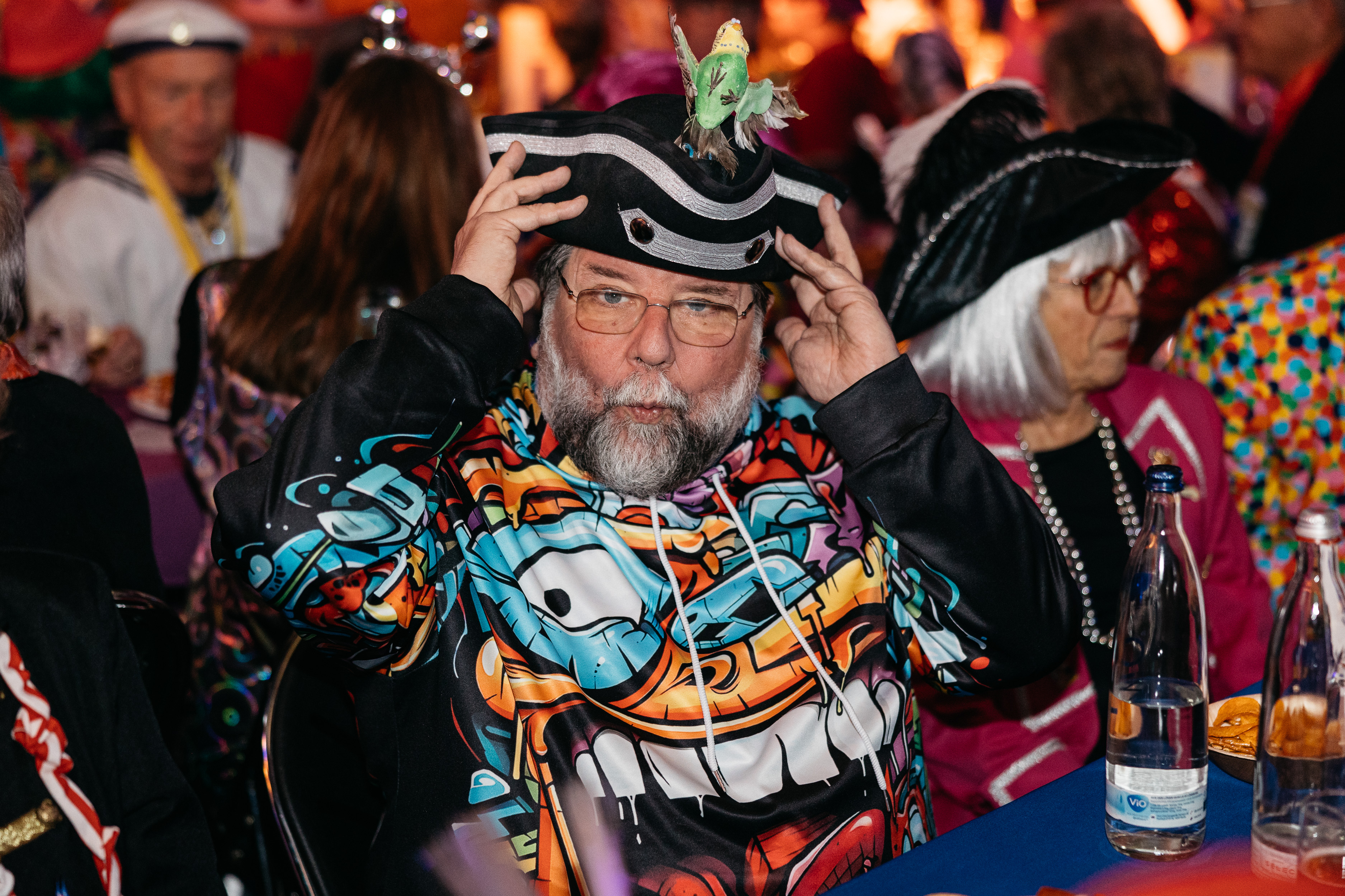 Mann mit grauem Bart trägt buntes Kostüm mit Comic-Motiv und Piratenhut mit grüner Figur darauf. Er sitzt an einem Tisch und blickt in die Kamera, im Hintergrund sind weitere kostümierte Personen zu sehen. 