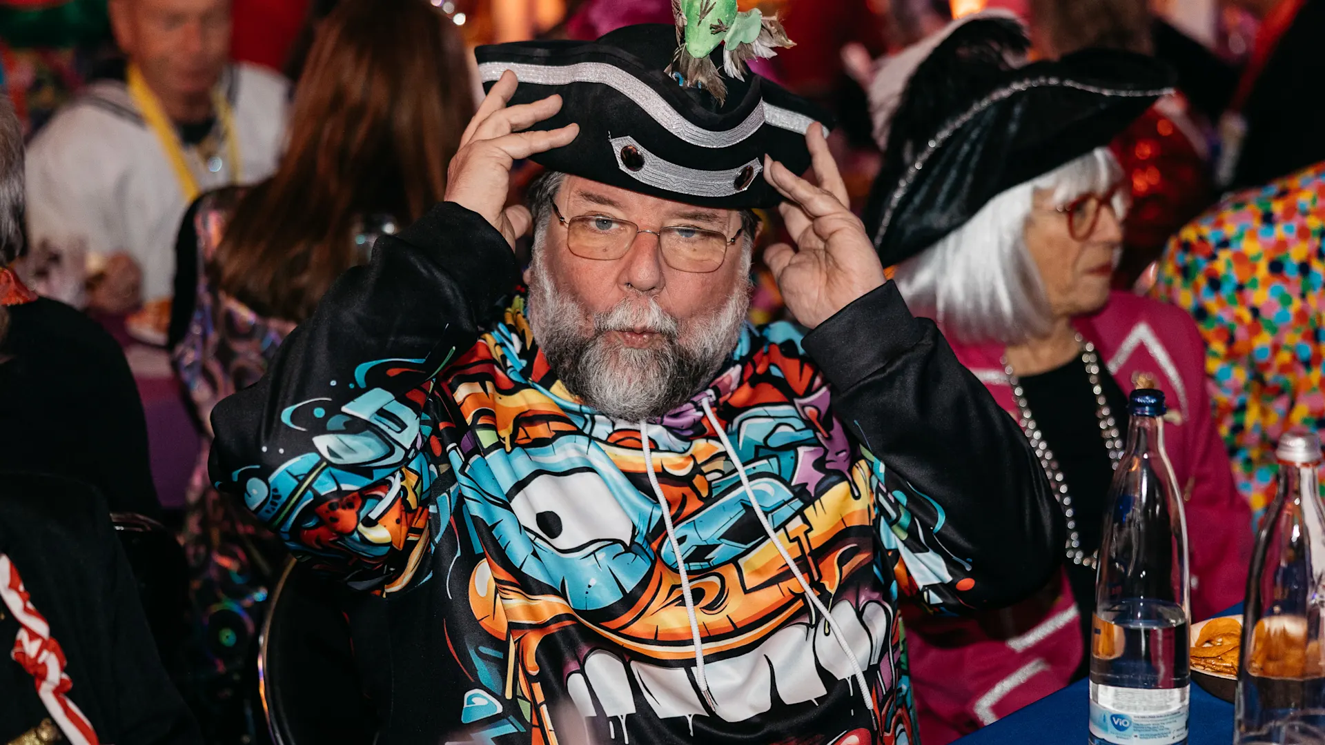 Mann mit grauem Bart trägt buntes Kostüm mit Comic-Motiv und Piratenhut mit grüner Figur darauf. Er sitzt an einem Tisch und blickt in die Kamera, im Hintergrund sind weitere kostümierte Personen zu sehen. Mann mit grauem Bart trägt buntes Kostüm mit Comic-Motiv und Piratenhut mit grüner Figur darauf. Er sitzt an einem Tisch und blickt in die Kamera, im Hintergrund sind weitere kostümierte Personen zu sehen.