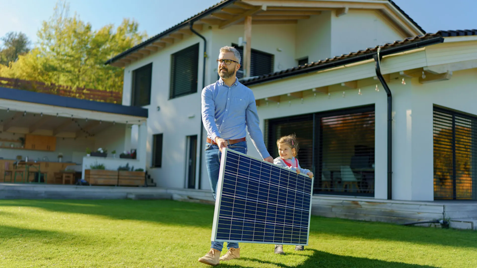 Ziele setzen, Emissionen senken. Ein Mann in blauem Hemd hält zusammen mit einem Kind ein Solarmodul in den Händen und läuft damit über eine Wiese. Das Haus hinter ihnen hat eine helle Fassade und Fenster die große Teile der Fassade einnehmen.