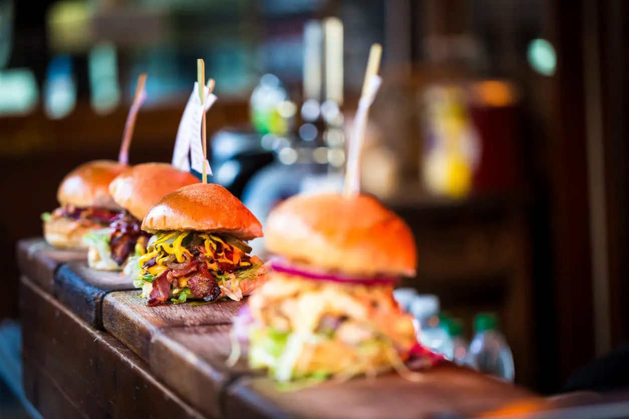 Nahaufnahme einer Auswahl frisch gegrillter Burger in Reihe auf einer Holztheke, Jeder der Burger hat sein eigenes Etikett, auf dem der Inhalt des Burgers steht. Die Burger werden zwischen glasierten Brötchen eingeklemmt und auf Betten mit frischem grünem Salat präsentiert und mit geschmolzenem Käse und roten Zwiebeln gefüllt. (Copyright coldsnowstorm Stock-Fotografie-ID:914886270)