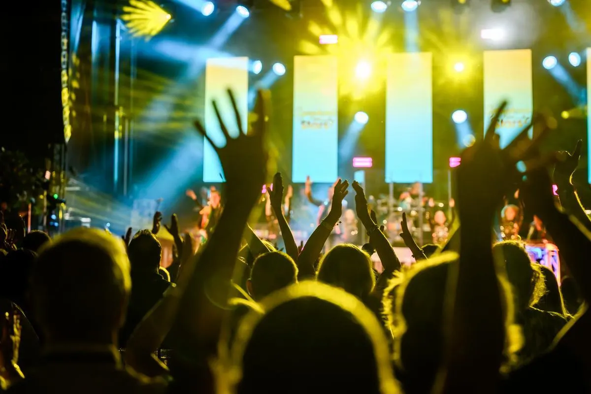 Lebendige Konzert- oder Veranstaltungsszene mit begeistertem Publikum. Zahlreiche Menschen strecken ihre Hände in die Luft, während Bühnenscheinwerfer in blau und gelb die Atmosphäre erhellen. Im Hintergrund ist eine Bühne mit Musiker:innen zu erkennen. 