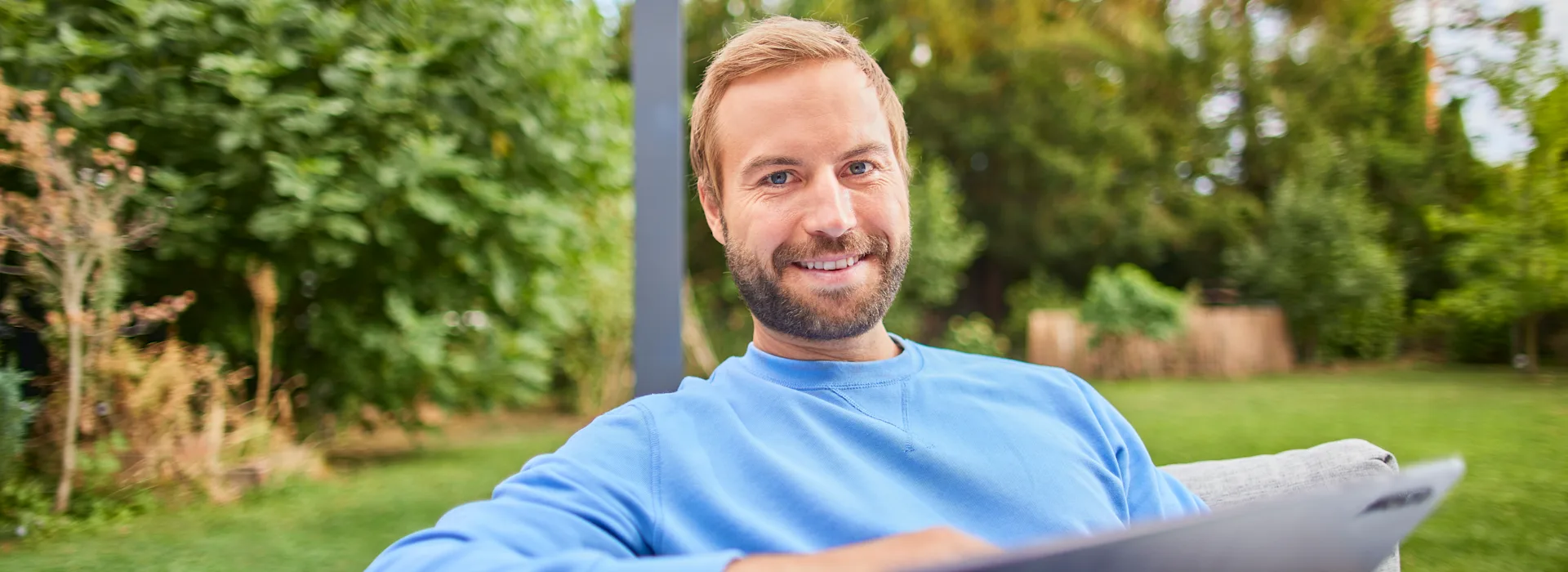 NEW Services entdecken Ein Mann sitzt mit einem Tablet im Garten und entdeckt die Tarife und Services der NEW.