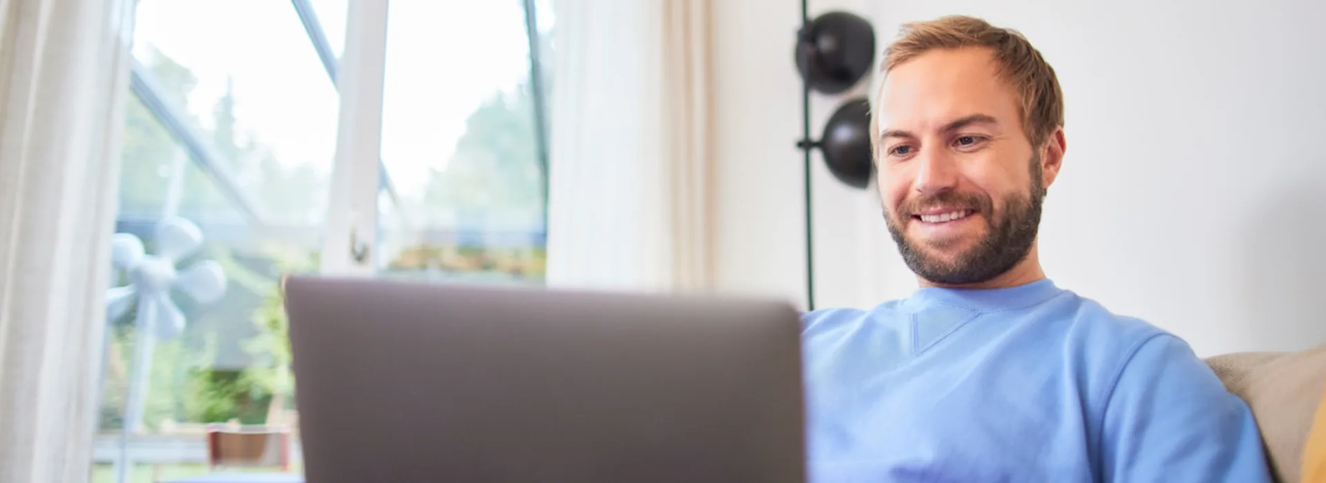 NEW-AG-Alles-zur-Bewerbung-H8A4604 Ein Mann sitzt bei sich Zuhause auf dem Sofa und schaut auf seinen Laptop.