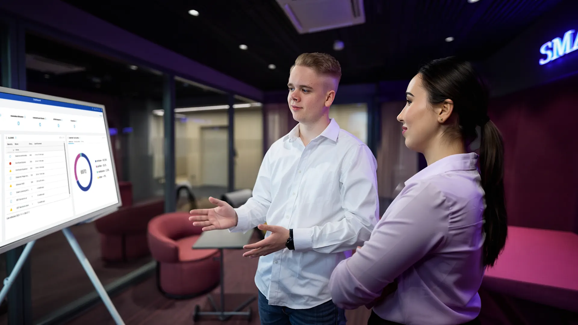 Zwei Azubis stehen vor einem Monitor Ein junger Mann in hellblauem Hemd erklärt einer Kollegin in weißer Bluse etwas auf einem großen Bildschirm mit Diagrammen und Datenvisualisierungen. Die beiden stehen in einem modernen Büroraum mit violetter und pinker Beleuchtung, roten Sitzmöbeln und Glaswänden.