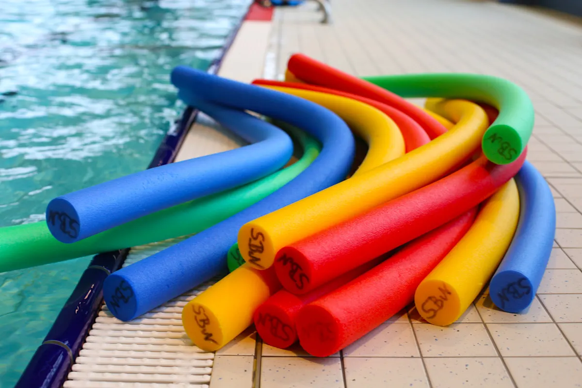 Auf dem Bild sieht man einen Stapel bunter Poolnudeln, die am Rand eines Schwimmbeckens liegen. Die Poolnudeln sind in verschiedenen Farben wie Blau, Grün, Gelb und Rot. Sie liegen auf einer gefliesten Fläche neben dem Wasser.
