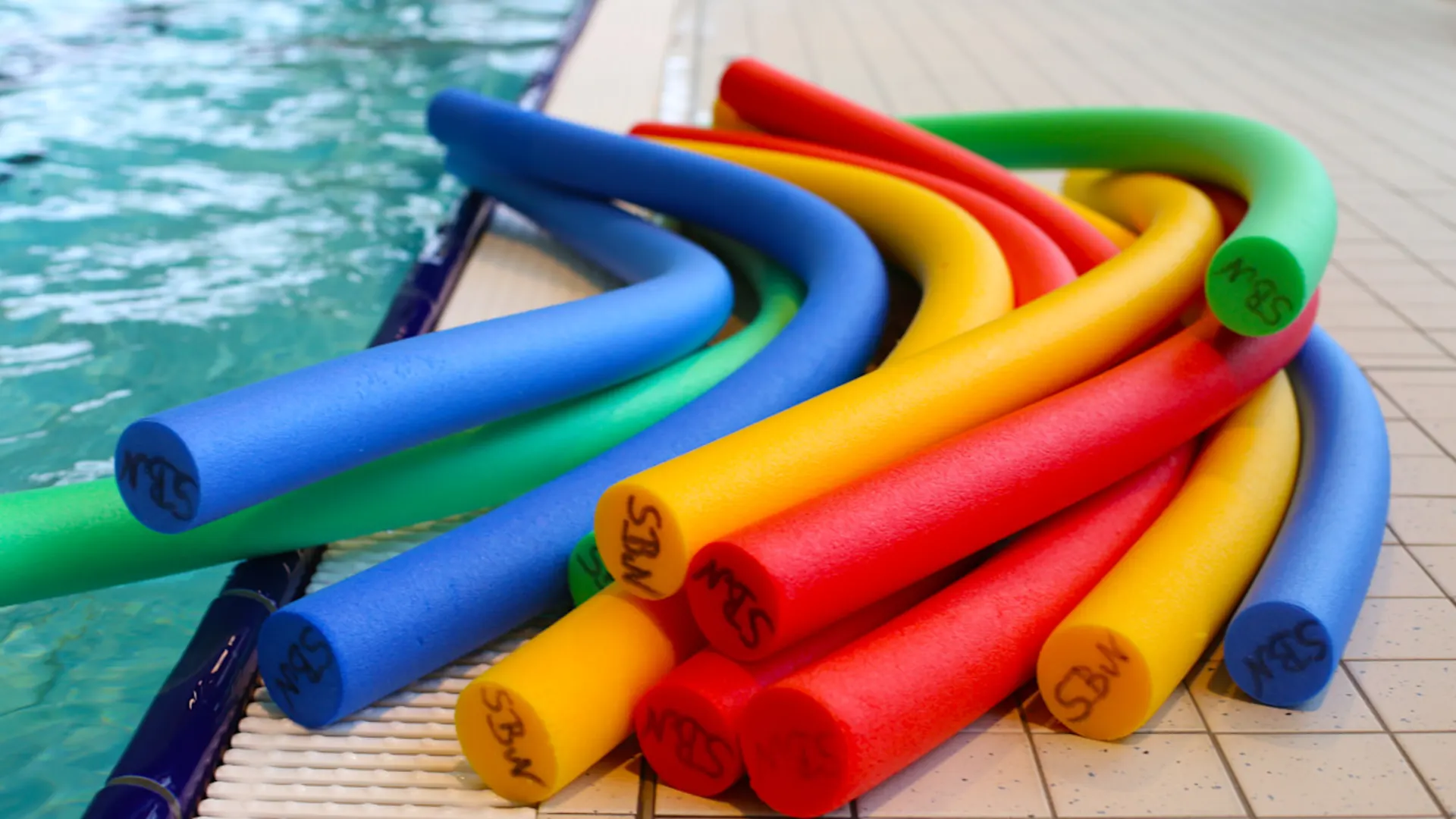 Schwimmhilfen am Beckenrand Auf dem Bild sieht man einen Stapel bunter Poolnudeln, die am Rand eines Schwimmbeckens liegen. Die Poolnudeln sind in verschiedenen Farben wie Blau, Grün, Gelb und Rot. Sie liegen auf einer gefliesten Fläche neben dem Wasser.