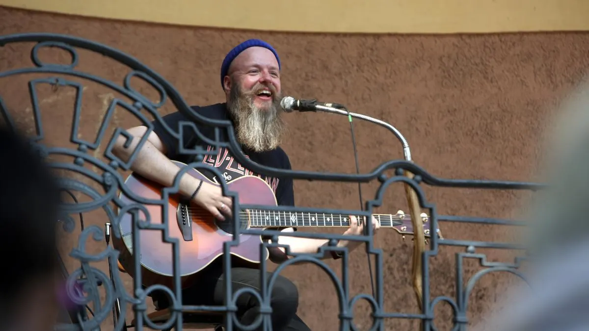 Ein Mann mit langem grauem Bart und blauer Mütze sitzt lächelnd mit einer Akustikgitarre vor einem Mikrofon auf einer Bühne. Er trägt ein schwarzes T-Shirt und performt. Vor ihm befindet sich ein Metallgeländer. 