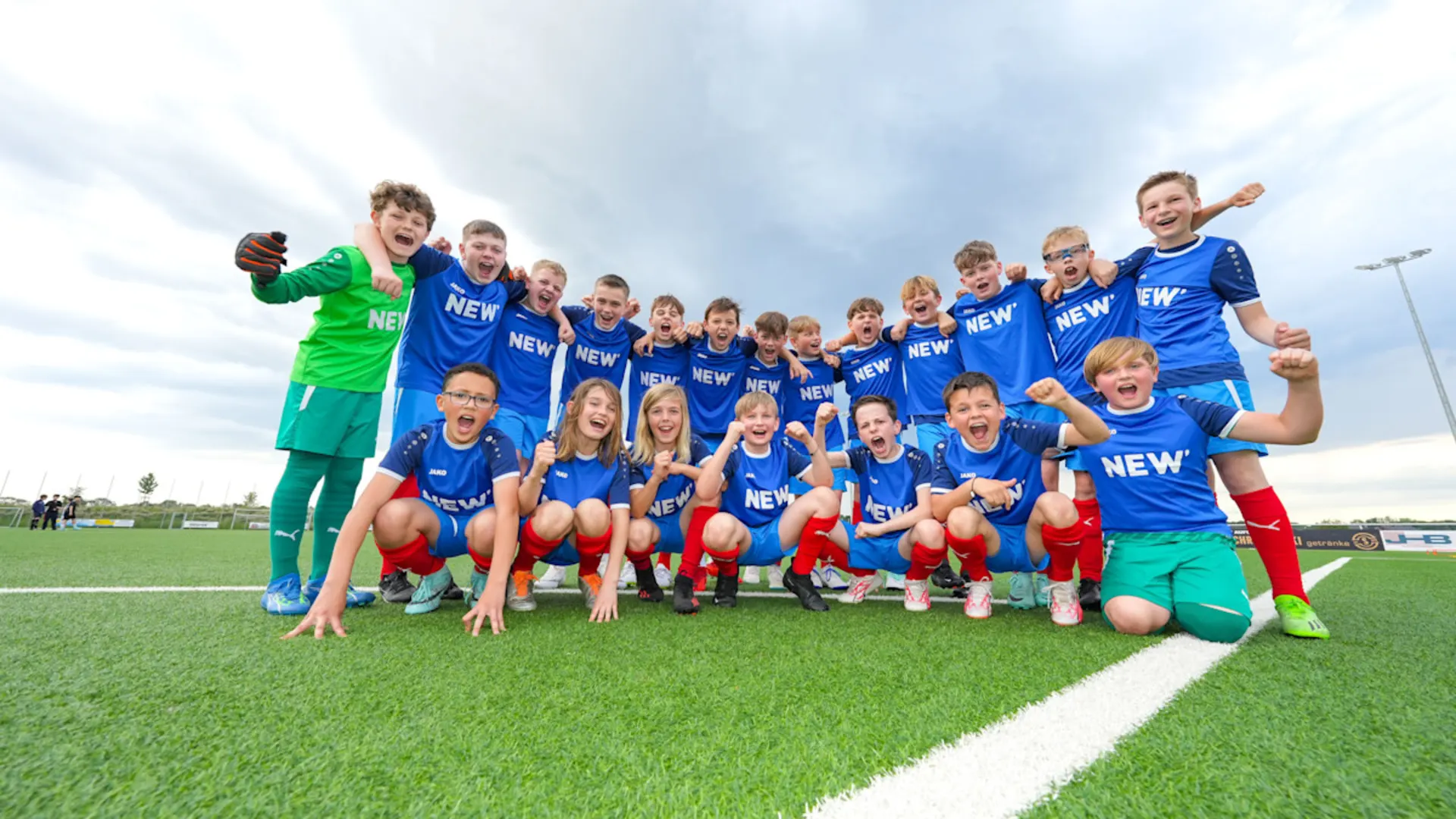 Menschen verbinden, Gemeinschaft stärken. Eine Fußballmannschaft in blauen Trikots und roten Hosen posiert gemeinsam für ein Gruppenfoto auf einem grünen Fußballplatz. Die Spieler sind in zwei Reihen aufgestellt, wobei einige knien und andere stehen, unter einem bewölkten Himmel.