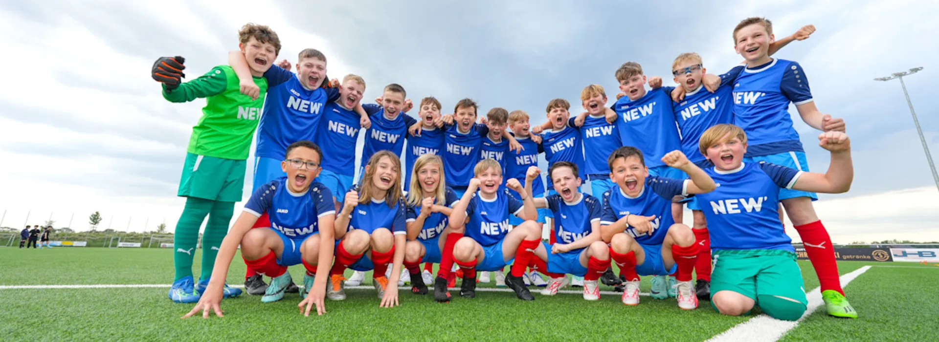 Menschen verbinden, Gemeinschaft stärken. Eine Fußballmannschaft in blauen Trikots und roten Hosen posiert gemeinsam für ein Gruppenfoto auf einem grünen Fußballplatz. Die Spieler sind in zwei Reihen aufgestellt, wobei einige knien und andere stehen, unter einem bewölkten Himmel.