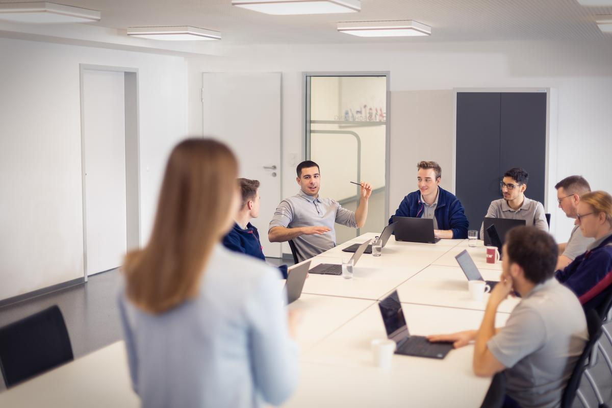 Eine Frau mit langen braunen Haaren steht vor einer Gruppe in einem modernen Konferenzraum. Weitere Personen sitzen um einen hellen Konferenztisch mit Laptops. Eine Person am Tisch erzählt etwas, alle Blicke sind auf diese Person gerichtet. 