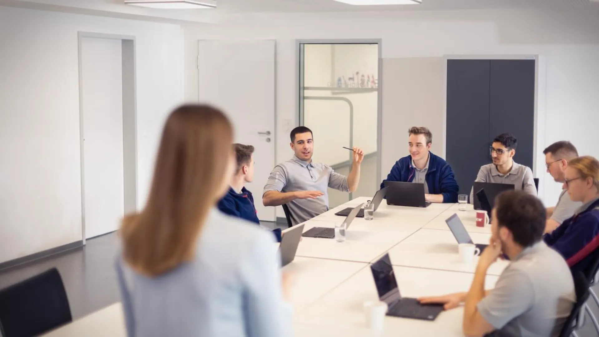Strategisch handeln, nachhaltig wirken. Eine Frau mit langen braunen Haaren steht vor einer Gruppe in einem modernen Konferenzraum. Weitere Personen sitzen um einen hellen Konferenztisch mit Laptops. Eine Person am Tisch erzählt etwas, alle Blicke sind auf diese Person gerichtet.