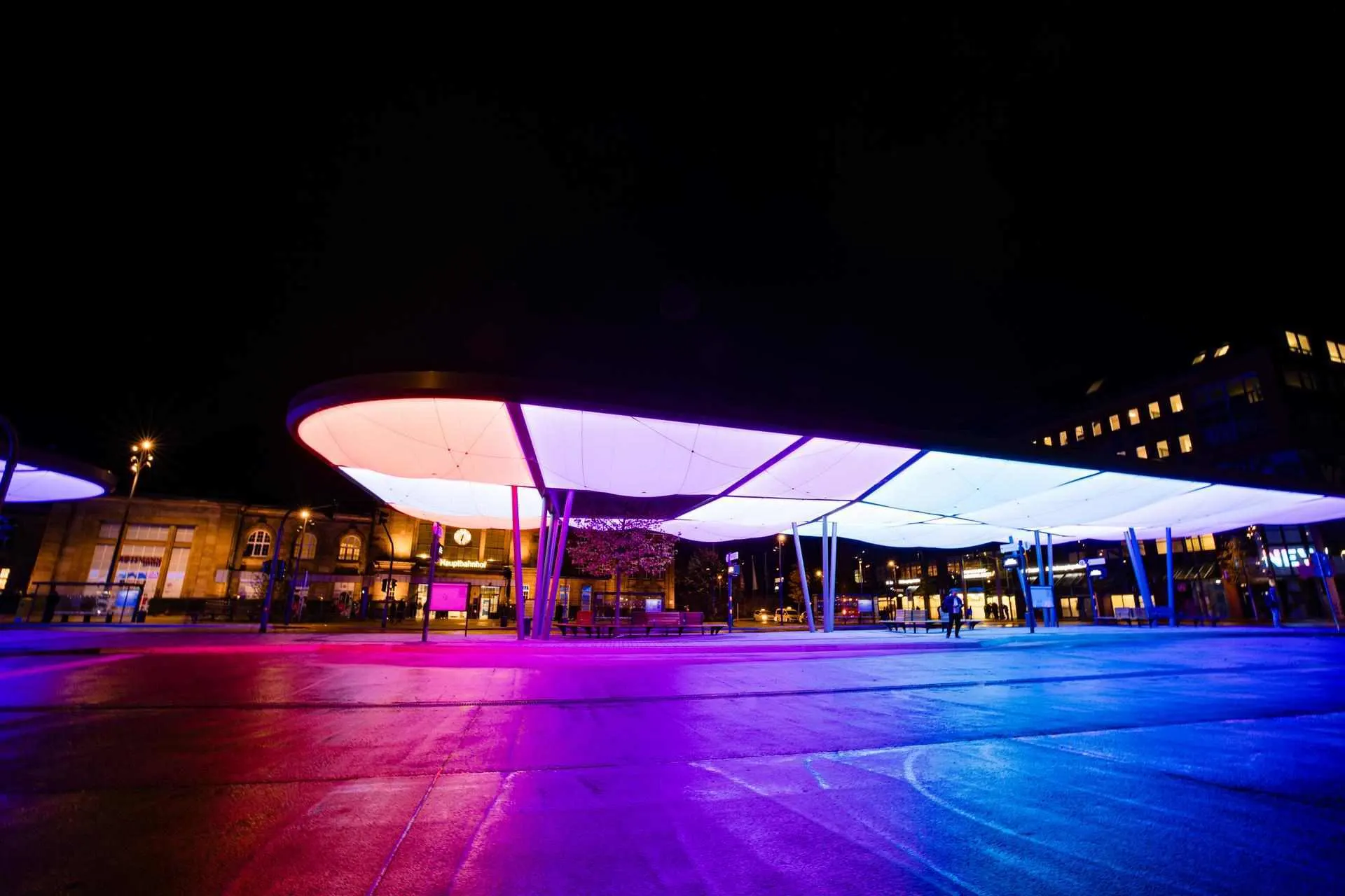 Das Bild zeigt den Zentralen Omnibusbahnhof (ZOB) in Mönchengladbach bei Nacht. Die moderne Dachkonstruktion ist farbig beleuchtet und prägt das Stadtbild. Im Hintergrund befindet sich der Hauptbahnhof