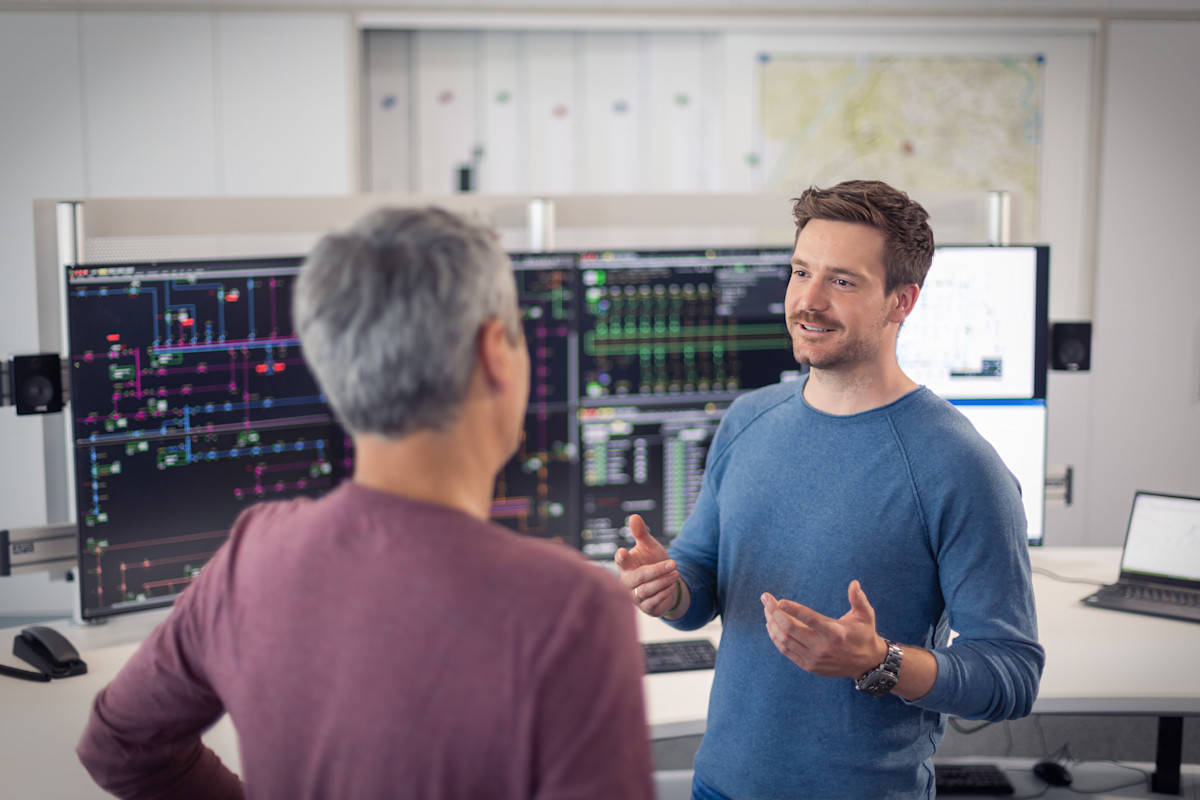 Zwei Kollegen führen ein Gespräch in einem Büroraum. Ein jüngerer Mann in blauem Pullover gestikuliert erklärend, während ein älterer Kollege in bordeauxrotem Shirt ihm zugewandt steht. Im Hintergrund sind mehrere große Monitore mit technischen Übersichtsdarstellungen und Kontrollsystemen zu sehen.