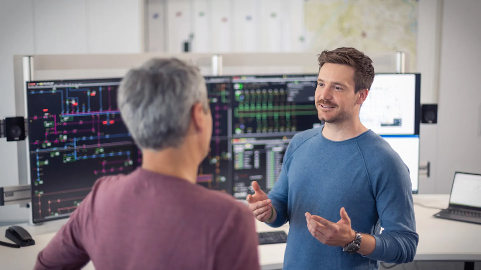 Veränderung gestalten, zukunftsfähig bleiben. Zwei Kollegen führen ein Gespräch in einem Büroraum. Ein jüngerer Mann in blauem Pullover gestikuliert erklärend, während ein älterer Kollege in bordeauxrotem Shirt ihm zugewandt steht. Im Hintergrund sind mehrere große Monitore mit technischen Übersichtsdarstellungen und Kontrollsystemen zu sehen.