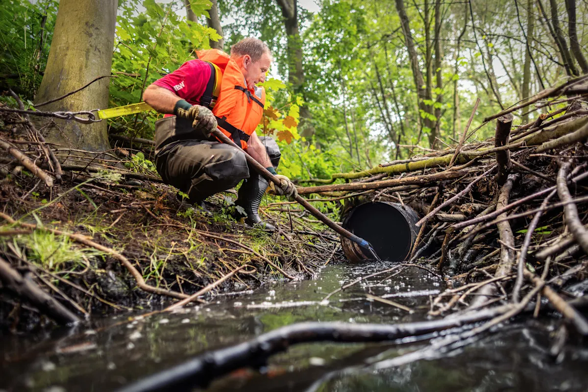 Ein Mann mit einer orangenen Weste hockt im Wald an einem Fluss, wo viele Äste von Bäumen drumherum liegen. Er ist hinten mit einem Karabinerhaken befestigt und fischt mit einem Stock in dem Fluss.
