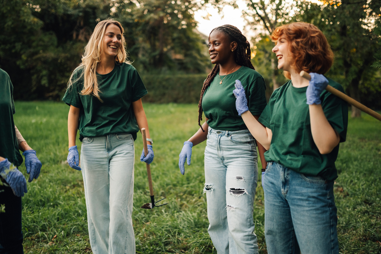 Das Bild zeigt drei junge Frauen auf einer Grünfläche, die an einer Freiwilligenarbeit oder einem Umweltprojekt teilnehmen. Alle tragen einheitliche grüne T-Shirts und hellblaue Jeans sowie blaue Arbeitshandschuhe. Die Frauen lächeln im Gespräch miteinander.