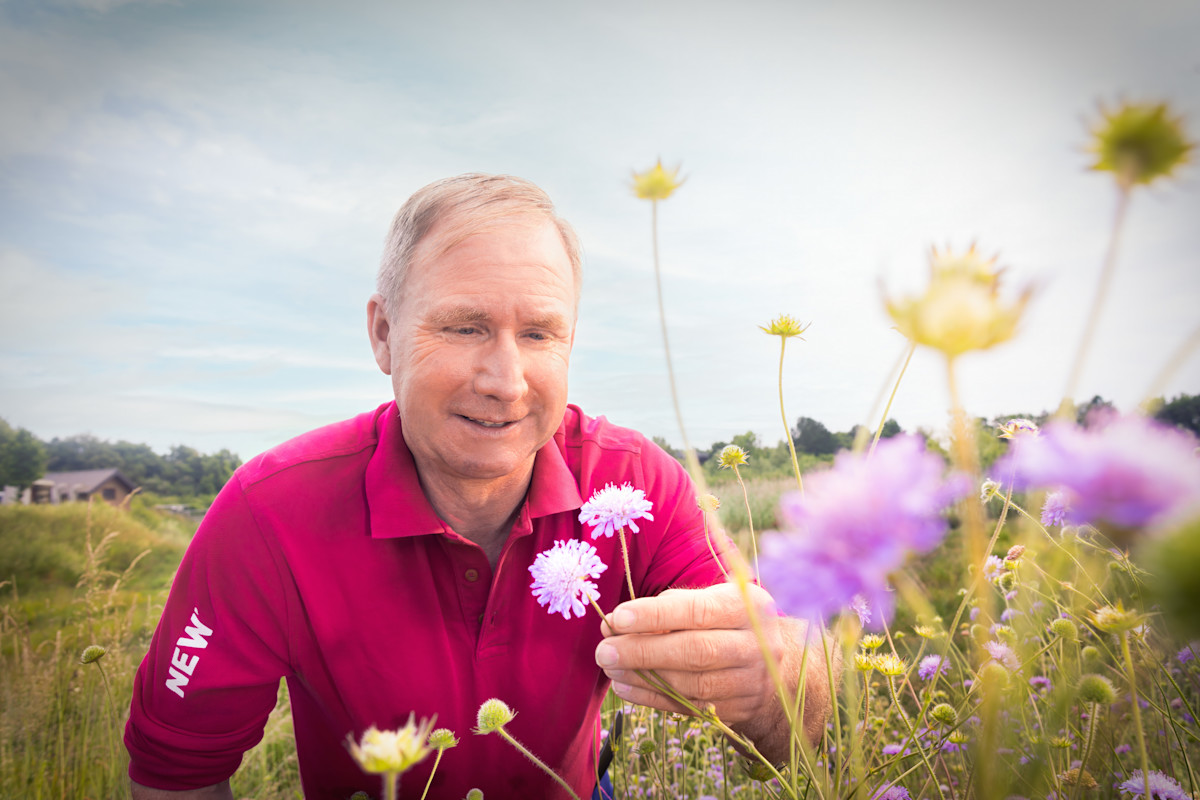 Mann im NEW Poloshirt auf einer Wildwiese mit einer Blume in der Hand.