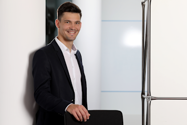 Ein junger Mann in schwarzem Anzug und weißem Hemd steht lächelnd an einer Glaswand in einem modernen Bürogebäude. Er hat dunkles, kurzes Haar und posiert professionell für ein Businessfoto. Ein junger Mann in schwarzem Anzug und weißem Hemd steht lächelnd an einer Glaswand in einem modernen Bürogebäude. Er hat dunkles, kurzes Haar und posiert professionell für ein Businessfoto.