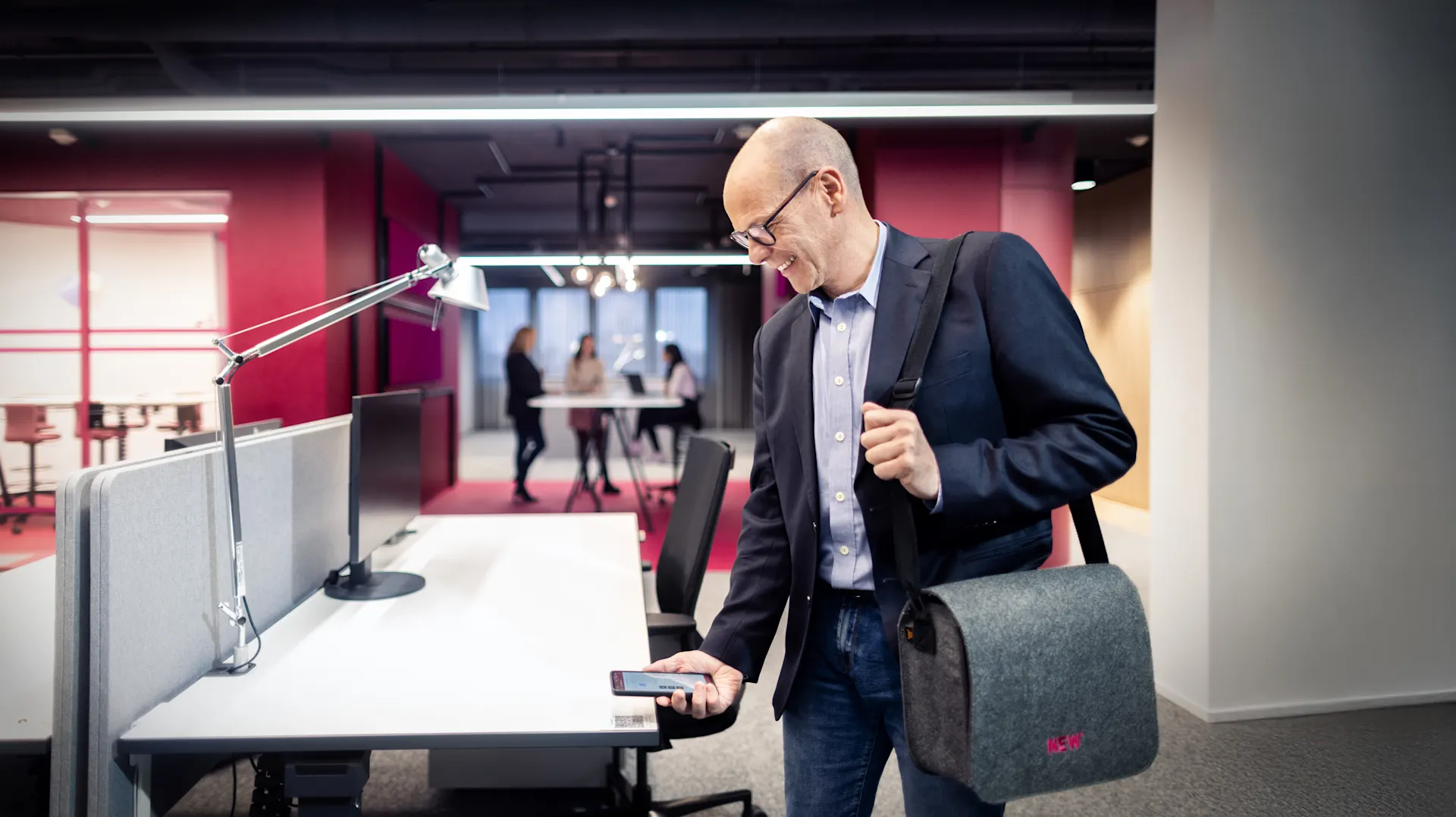 Mitarbeiter der NEW AG meldet seinen Schreibtisch per QR Code ab.  Er trägt eine Laptop-Tasche und hat sein Smartphone in der Hand, im Hintergrund stehen Personen an einem Tisch in offener Arbeitsumgebung.
