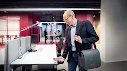 Mitarbeiter der NEW AG meldet seinen Schreibtisch per QR Code ab.  Er trägt eine Laptop-Tasche und hat sein Smartphone in der Hand, im Hintergrund stehen Personen an einem Tisch in offener Arbeitsumgebung.