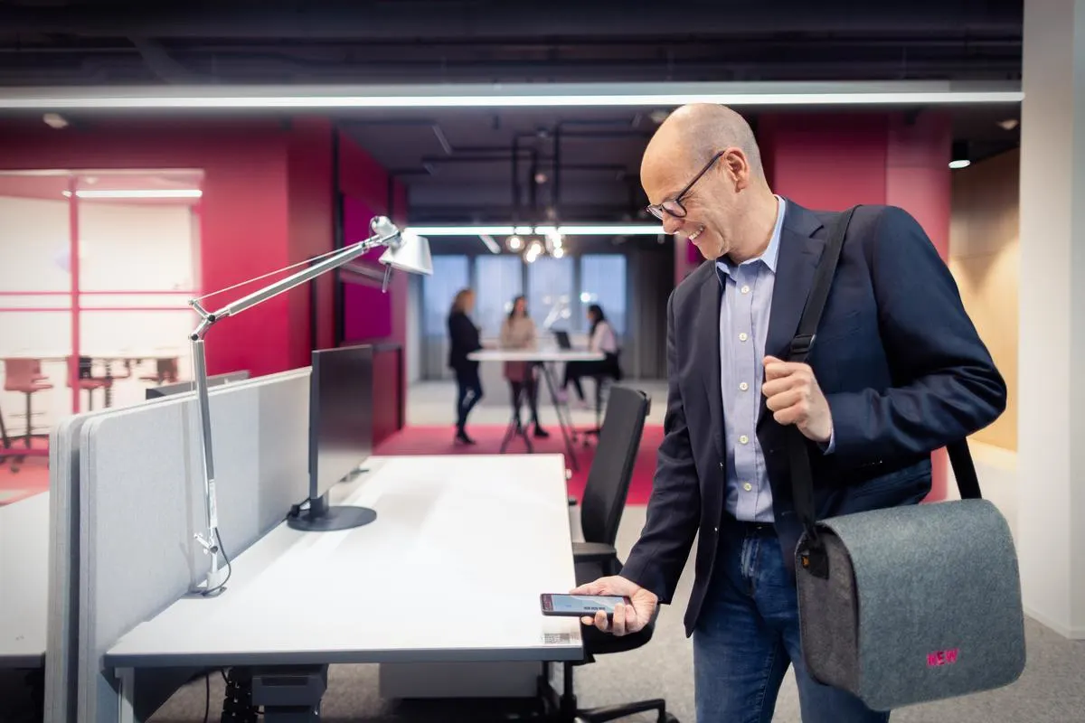 Mitarbeiter der NEW AG betritt ein modernes Büro mit Laptop-Tasche und Smartphone in der Hand, im Hintergrund stehen Personen an einem Tisch in offener Arbeitsumgebung.