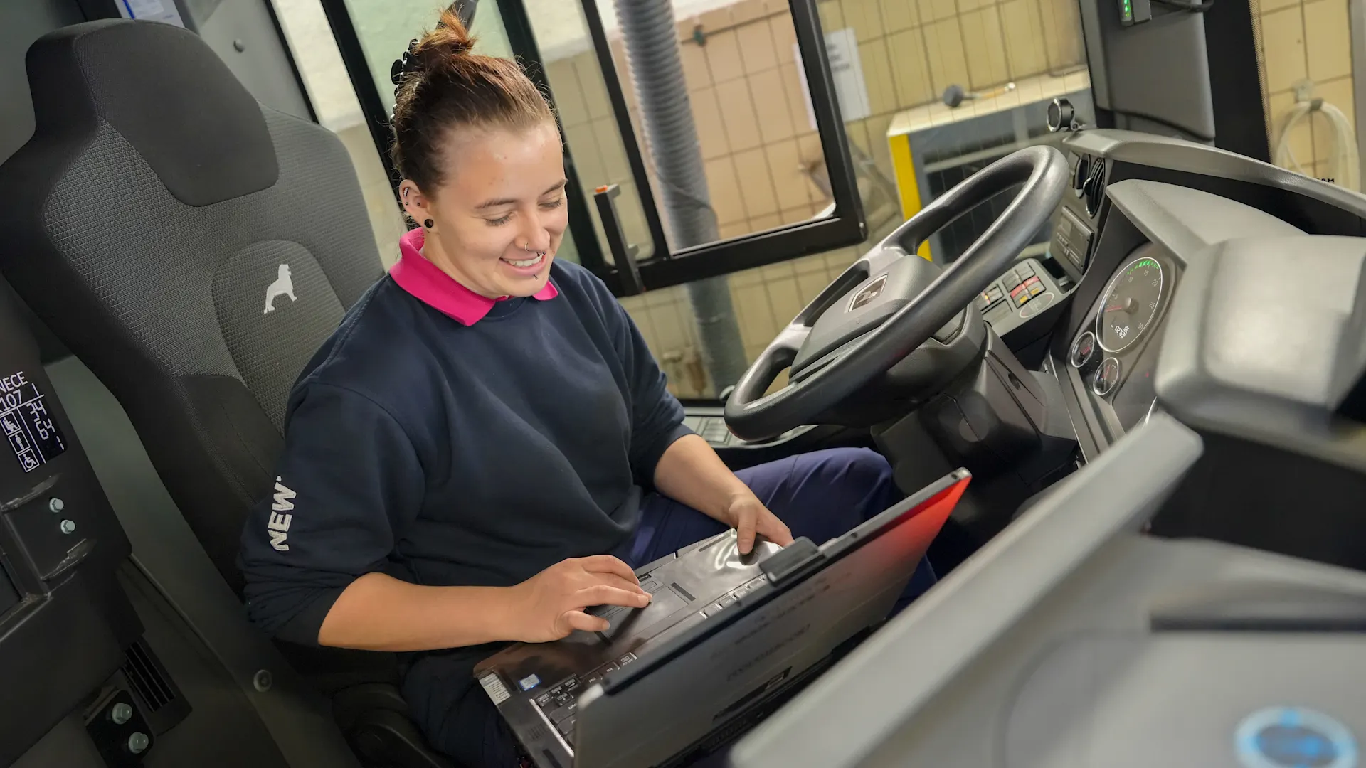 Kfz-Mechatronikerin Daniela Albers Daniela Albers, Auszubildende zur Kfz-Mechatronikerin, sitzt auf dem Fahrersitz eines stehenden Fahrzeugs und arbeitet an einem Laptop.