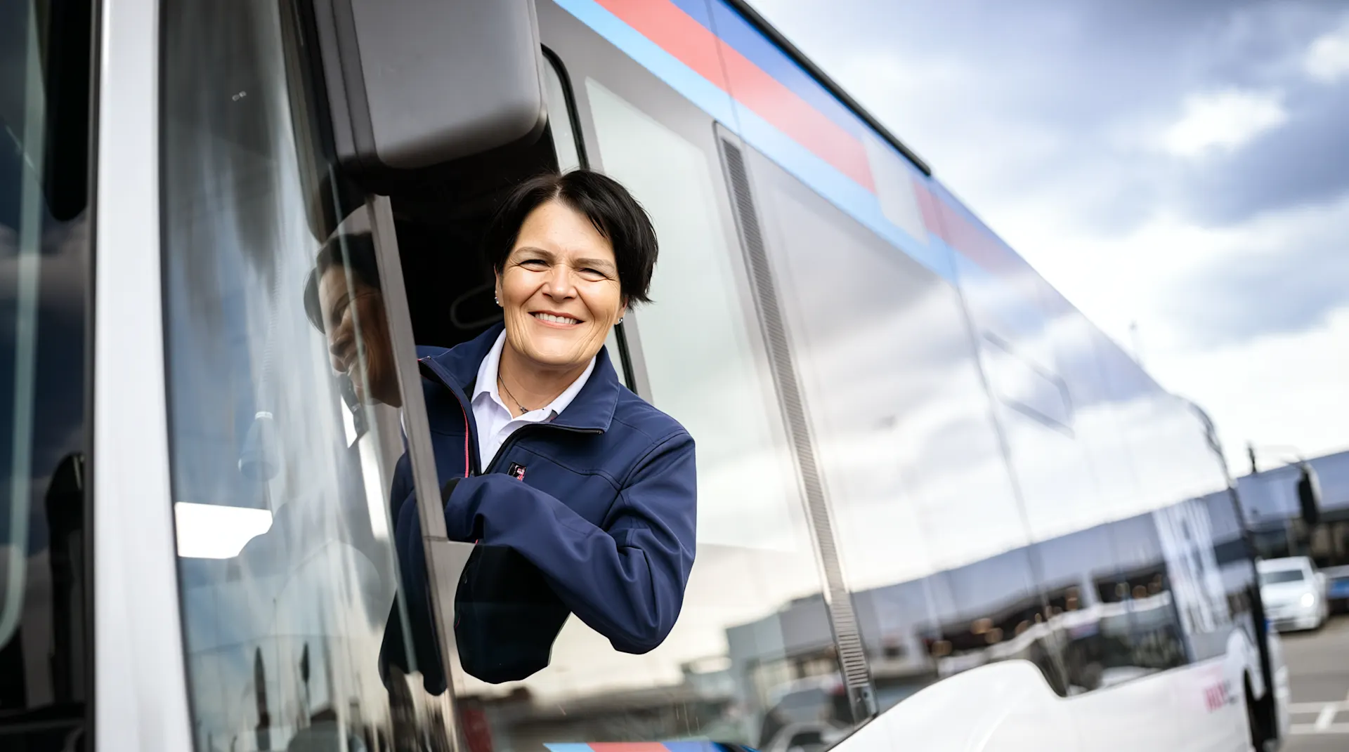 Eine lächelnde Busfahrerin in dunkelblauer Arbeitsjacke lehnt sich aus dem geöffneten Fenster ihres Busses. Im Hintergrund ist die moderne Busfassade mit türkis-rosa Streifen und ein bewölkter Himmel zu sehen.