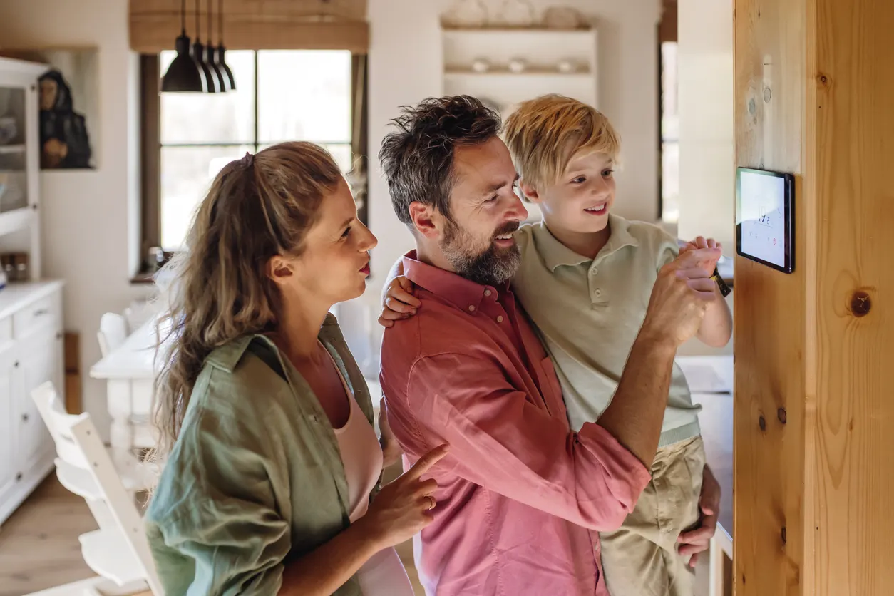 Eine junge Familie steht vor einem Smart-Home-Panel an der Wand. Der Vater hält seinen Sohn auf dem Arm, während der Junge auf das Bedienfeld tippt. Die Mutter schaut auf das Display. 