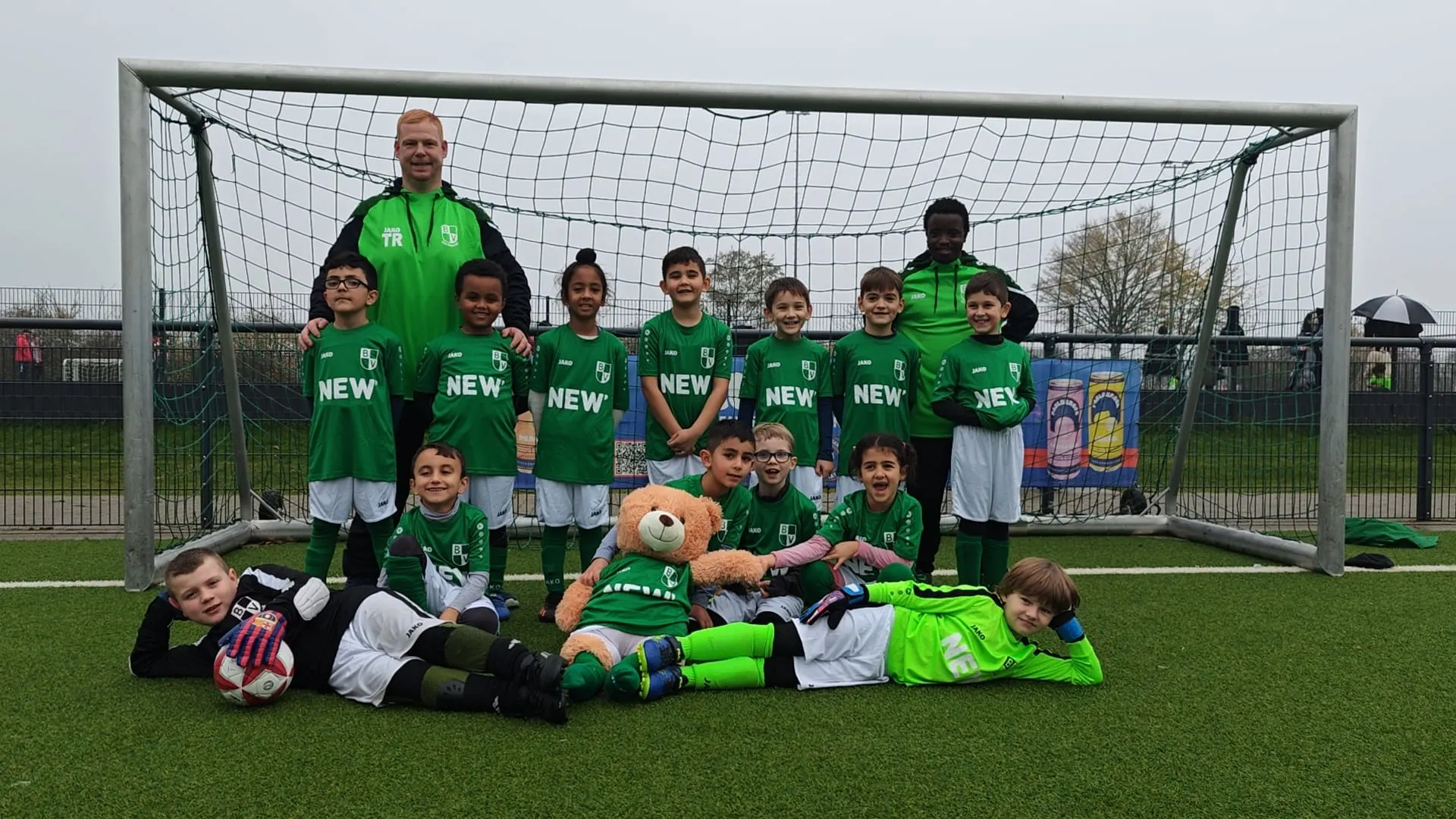 Martin als Fußballtrainer der F-Jugend. Martin Giesers-Aviles steht als Trainer in grünem Trainingsanzug hinter seiner F-Jugend-Fußballmannschaft, die in grünen NEW-Trikots vor einem Fußballtor auf dem Kunstrasenplatz posiert, einige Kinder stehen, andere liegen vorne mit Bällen und einem Maskottchen.